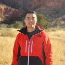 Man in red jacket smiling in dry grass with red rock cliffs behind (photo)